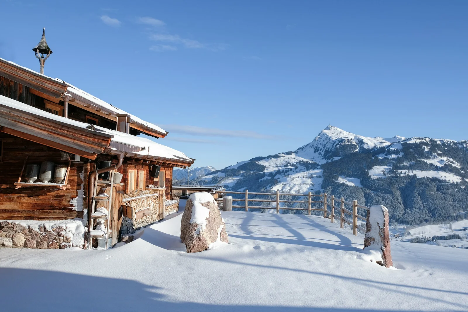 Chalet en bois à Kitzbühel, dans les Alpes autrichiennes, sous le soleil, entouré de neige face aux sommets.