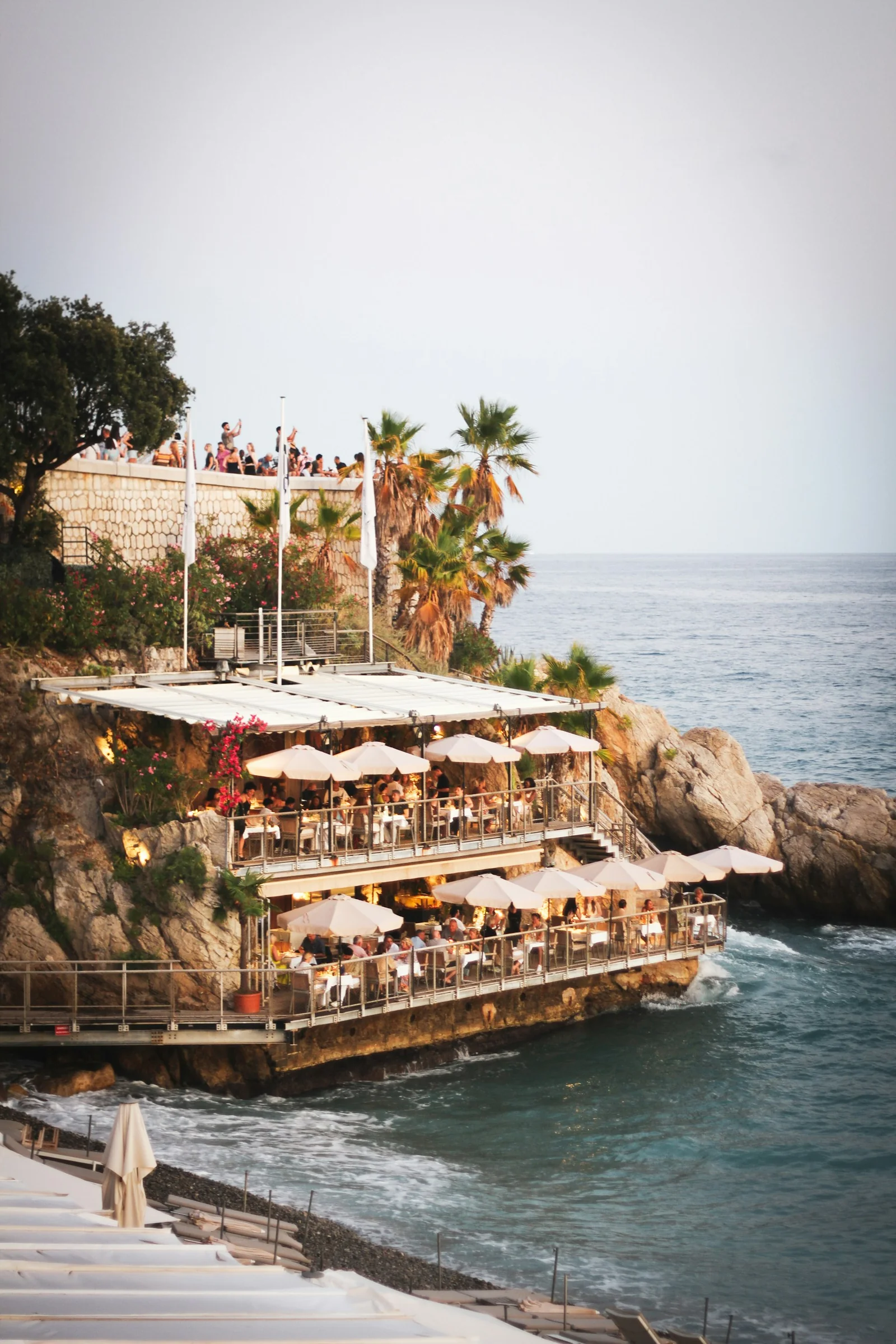 Terrasse de restaurant au-dessus de la mer à Nice, tables sous des parasols au bord de la falaise.