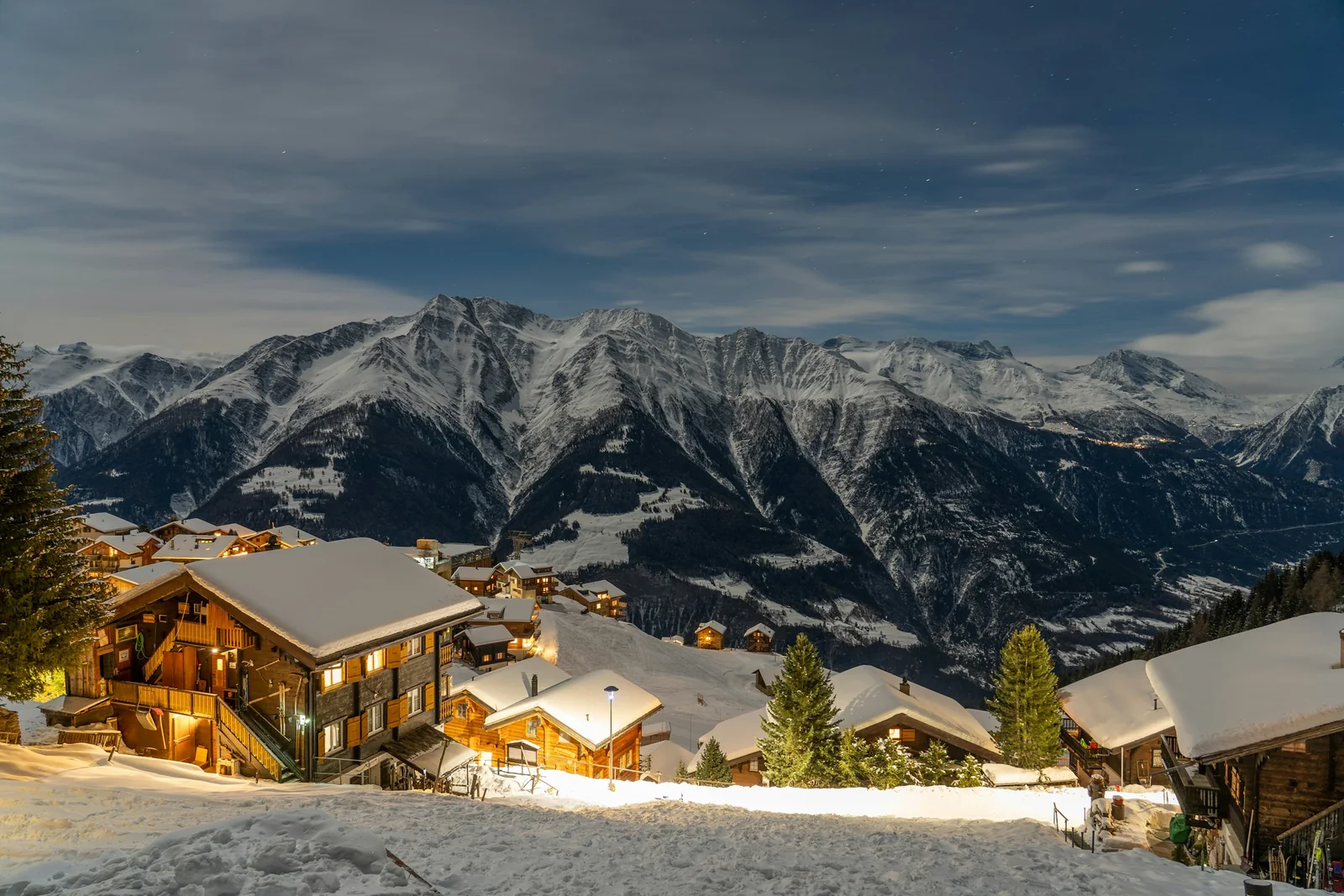 Chalets enneigés à Riederalp, fenêtres éclairées de lumière chaude face aux sommets des Alpes sous un ciel bleu nocturne.